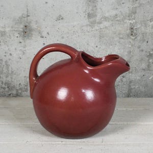 May include: A round, deep red ceramic pitcher with a smooth, glossy finish. The vessel features a curved handle on the left side and a wide, integrated pouring spout on the right, showcasing a simple, bulbous silhouette and uniform color.