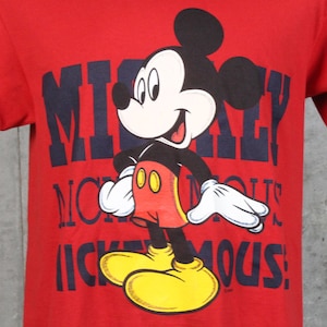 May include: Red t-shirt featuring a graphic of Mickey Mouse. The cartoon character is in a standing pose with a smiling expression. The shirt has the words "Mickey Mouse" in navy blue block letters.