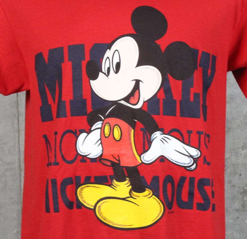 May include: Red t-shirt featuring a graphic of Mickey Mouse. The cartoon character is in a standing pose with a smiling expression. The shirt has the words "Mickey Mouse" in navy blue block letters.