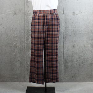 May include: Plaid pants in a straight-leg style. The pants feature a classic plaid pattern in shades of navy blue, brown, and orange. The pants are displayed on a stand against a gray concrete wall. The pants have two front pockets.