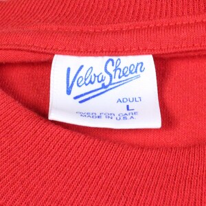 May include: Close-up of a red t-shirt with a white tag. The tag features the brand name "Velva Sheen" in blue script, with "ADULT L" and "OVER FOR CARE MADE IN U.S.A." printed below.
