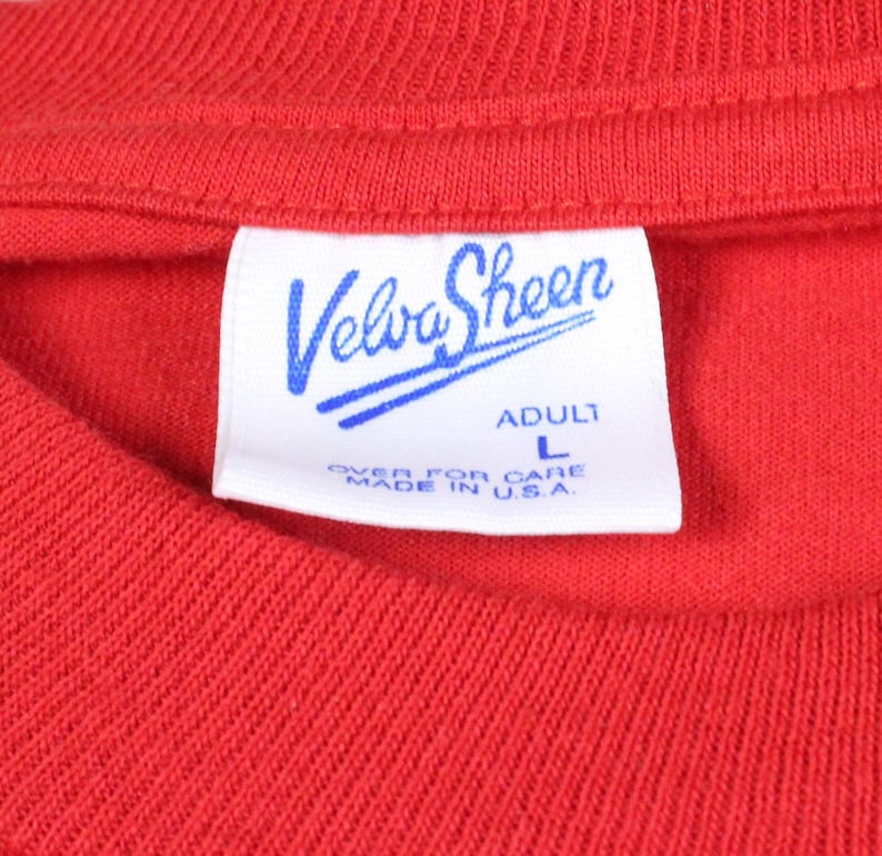 May include: Close-up of a red t-shirt with a white tag. The tag features the brand name "Velva Sheen" in blue script, with "ADULT L" and "OVER FOR CARE MADE IN U.S.A." printed below.