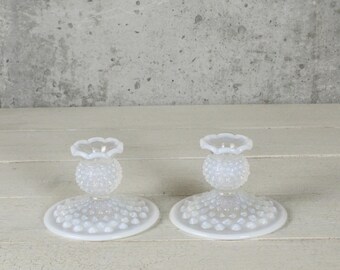 May include: Two white glass candle holders. Each has a round base with a textured pattern, a textured spherical center, and a scalloped top. The candle holders are displayed on a white wooden surface.