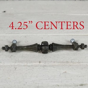 May include: A dark bronze-toned metal drawer pull with a decorative, elongated spindle shape and a central bulbous detail. The hardware features two mounting points with silver-colored screws and includes the text 4.25" CENTERS above it.