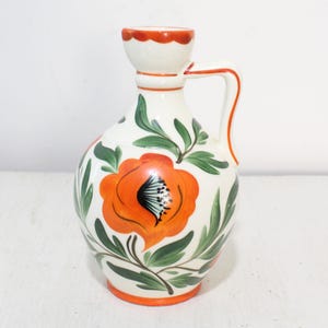 Może przedstawiać: Biały ceramiczny dzbanek z uchwytem, ozdobiony żywym pomarańczowym kwiatem i wzorem zielonych liści. Obręcz i podstawa są pomarańczowe. Ten dekoracyjny dzbanek jest idealny do serwowania napojów lub jako element dekoracyjny.