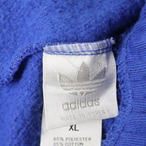 May include: A white tag with black text that reads "adidas" and "Made in Korea". The tag also has a white Adidas logo and the text "XL", "65% Polyester", and "35% Cotton".