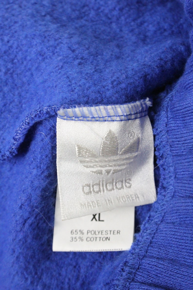 May include: A white tag with black text that reads "adidas" and "Made in Korea". The tag also has a white Adidas logo and the text "XL", "65% Polyester", and "35% Cotton".