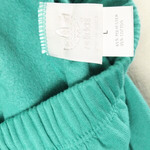 May include: A white tag with black text that reads "adidas" and "65% Polyester 35% Cotton" attached to a teal pair of sweatpants. The tag also has a size "L" printed on it.