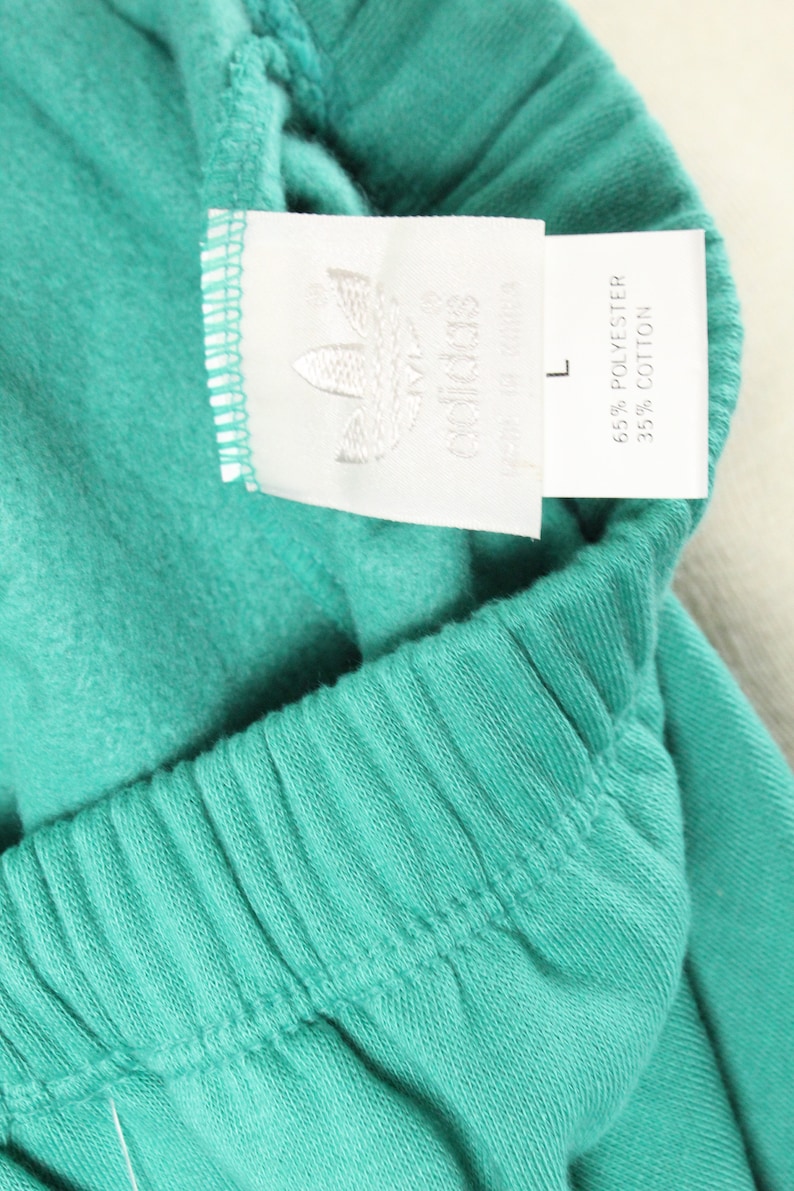 May include: A white tag with black text that reads "adidas" and "65% Polyester 35% Cotton" attached to a teal pair of sweatpants. The tag also has a size "L" printed on it.