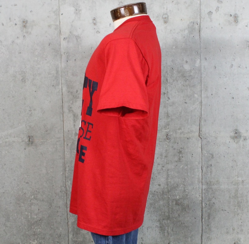 May include: A red t-shirt with navy blue lettering. The text on the shirt appears to read "CITY HOUSE" vertically. The shirt has short sleeves and a crew neck. The shirt is displayed on a mannequin.