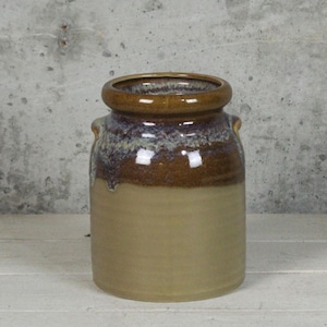 May include: A ceramic jar with a wide, rounded rim and two small handles. The top portion is a rich brown with a speckled, glazed finish, transitioning to a lighter tan color on the lower half. The jar has a rustic, handcrafted appearance.