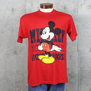 May include: Red t-shirt featuring a graphic of Mickey Mouse. The design includes Mickey Mouse in his classic outfit with yellow shoes, red shorts, and white gloves. The text "Mickey Mouse" is printed in navy blue.