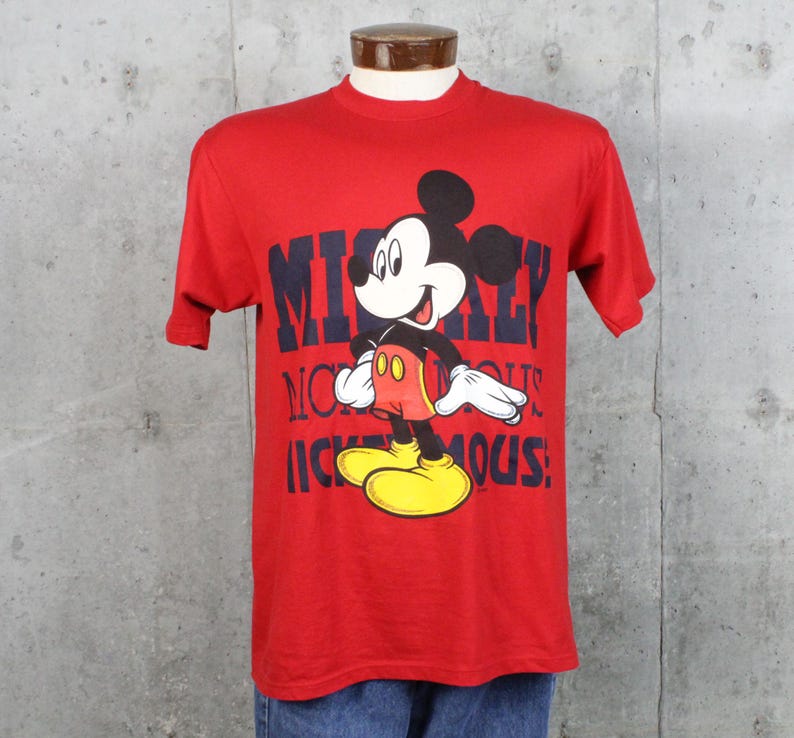 May include: Red t-shirt featuring a graphic of Mickey Mouse. The design includes Mickey Mouse in his classic outfit with yellow shoes, red shorts, and white gloves. The text "Mickey Mouse" is printed in navy blue.