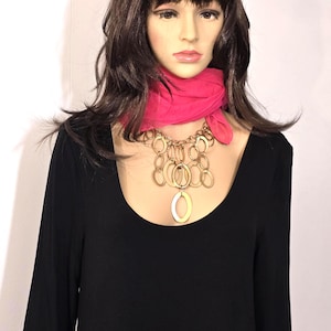 May include: A black long-sleeve dress with a scoop neckline, paired with a gold-toned necklace featuring multiple oval pendants. A bright pink scarf is draped around the neck. The mannequin has brown hair.