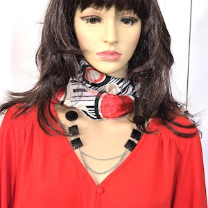 May include: A mannequin wearing a red long-sleeved blouse with a V-neck and button details. A black and silver necklace is worn over the blouse. A patterned scarf with red, black, and white designs is around the neck.