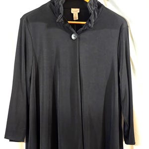 May include: A black long-sleeve top with a high, ruffled collar and a single button closure. The top is hanging on a wooden hanger. The fabric appears to be a soft, flowing material.