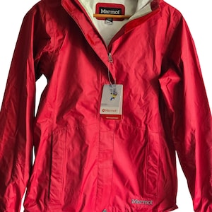 Könnte beinhalten: Eine leuchtend rote Regenjacke mit weißem Innenfutter, einem durchgehenden Frontreißverschluss und einer Kapuze. Die Jacke zeigt den Markennamen Marmot in Weiß auf der Brust und ein hängendes Produktetikett mit dem Marmot-Logo und Text.