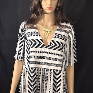 Peut inclure: Robe à motifs noir et blanc avec un col en V et des manches courtes. La robe présente des motifs géométriques et est accessoirisée d'un collier doré.