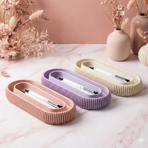 Può includere: Tre portapenne ovali nei colori pastello: rosa, lavanda e crema. Ogni portapenne ha un esterno a coste e contiene un vassoio interno abbinato con una penna bianca con punta nera. I portapenne sono disposti su una superficie marmorea.