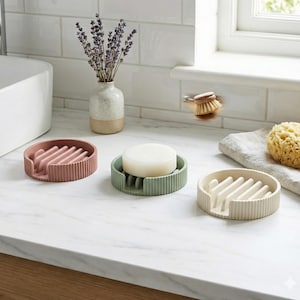 May include: Three round soap dishes in shades of pink, green, and cream, each with a ribbed design and a raised section for drainage. One dish holds a bar of soap. A vase of lavender and a natural sponge are also visible.