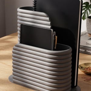 May include: A grey desk organiser with a ribbed, horizontal texture and two tiered compartments. It holds a large black spiral-bound notebook, a smaller black notebook, and a black pen with a gold clip, all resting on a flat oval base.