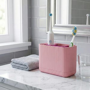 Puede incluir: Un soporte rosa para cepillos de dientes con líneas verticales, que contiene dos cepillos de dientes. Una toalla gris doblada y un vaso de agua están sobre la encimera de mármol. Se ven un cepillo de dientes eléctrico y uno manual.