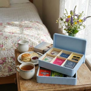 May include: A light blue tea box with vertical stripes, filled with tea bags, is open on a wooden table. A cup of tea, a scone with jam, and a small jug are nearby. A bouquet of flowers and a book are also on the table.
