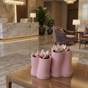 May include: Two pink, wavy-edged planters with faux succulents sit on a wooden table. The ribbed planters have a light pink hue and a unique, modern design. The background shows a lobby with marble, plants, and seating.