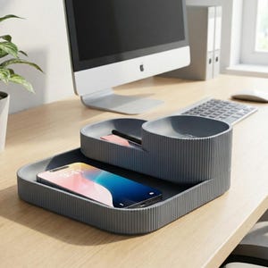 May include: A grey desk organiser with a ribbed design, holding a smartphone, pen, and small notepad. The phone displays a colourful screen. A computer monitor, keyboard, and mouse are in the background.
