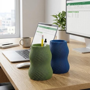 Puede incluir: Dos portalápices decorativos con un diseño ondulado y rayado. Uno es verde, el otro azul. Se ven lápices y bolígrafos en el portalápices verde. Los portalápices están sobre un escritorio de madera.