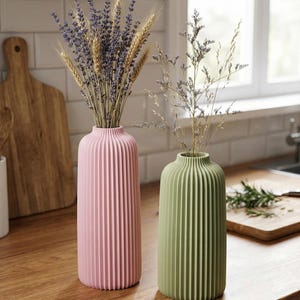 May include: Two decorative vases, one pink and one green, with vertical ribbed detailing. The pink vase holds lavender and wheat, while the green vase contains delicate dried flowers. The vases are on a wooden countertop.