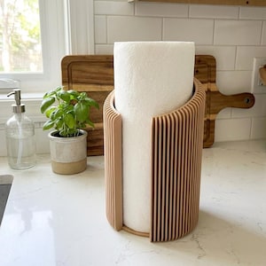 May include: A wooden paper towel holder with a roll of white paper towels. The holder has a vertical slat design in a light brown color. A small potted plant and a wooden cutting board are in the background.