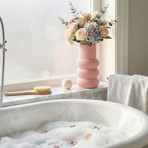 May include: A pink, ribbed ceramic vase holds a bouquet of pastel-colored flowers, including roses and hydrangeas. The vase sits on a marble ledge near a window, next to a bath brush and a bath bomb. A white bathtub is filled with bubbles and rose petals.