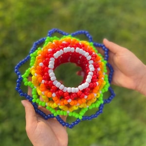 Puede incluir: Una colorida obra de arte con cuentas en un arcoíris de colores, incluyendo rojo, naranja, amarillo, verde y azul. El diseño circular es sostenido por manos sobre un fondo verde borroso. La pieza está hecha de pequeñas cuentas redondas.