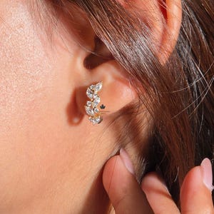 Könnte beinhalten: Nahaufnahme eines goldenen Ohrrings mit mehreren klaren Edelsteinen. Der Ohrring ist im kleinen Creolen-Stil gehalten, wobei die Edelsteine in einem gebogenen Muster angeordnet sind. Die Person hat braunes Haar.