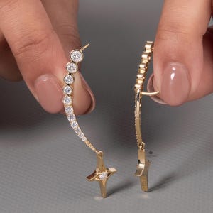 Könnte beinhalten: Ein Paar goldfarbene Ohrringe. Ein Ohrring hat ein geschwungenes Design mit einer Reihe kleiner, runder, klarer Steine und einem sternförmigen Anhänger. Der andere Ohrring hat ein ähnliches Design. Die Ohrringe werden vor einem grauen Hintergrund gehalten.