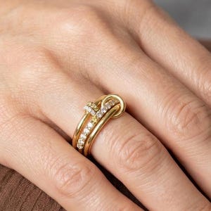 Könnte beinhalten: Ein Goldring mit mehrschichtigem Design und einem knotenartigen Mittelstück. Der Ring besteht aus einer glatten Goldschiene und zwei mit kleinen, klaren, funkelnden Steinen besetzten Schienen, die sich vorne überkreuzen und schichten.