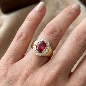 May include: A gold ring featuring a central oval-cut red gemstone surrounded by a halo of small round white stones. The band is smooth and polished, showcasing a classic design with a prominent setting that highlights the vibrant red centre stone.