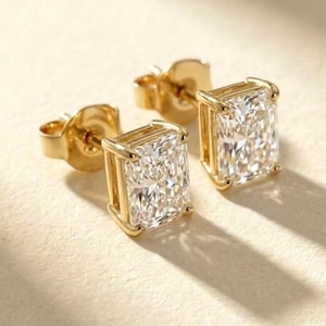 May include: A pair of gold stud earrings with rectangular, clear gemstones. The earrings are set in a gold frame, and the light reflects off the facets of the stones. The earrings are displayed on a light-colored surface.
