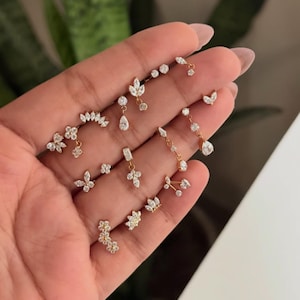 May include: A collection of gold-tone stud earrings featuring clear, sparkling cubic zirconia stones in various shapes, including floral, leaf, teardrop, and butterfly designs, arranged in a cluster on a palm for display.