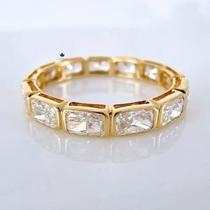 Könnte beinhalten: Ein goldener Bandring mit einer Reihe rechteckiger, klarer Edelsteine. Der Ring hat eine polierte, reflektierende Oberfläche, und die Edelsteine sind in einzelnen Fassungen gefasst. Der Ring wird auf einer weißen Oberfläche präsentiert.