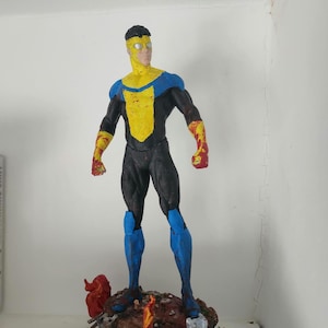 Op de afbeelding: Een beeldje van een superheld in een zwart, geel en blauw kostuum, staand op een rotsachtige basis met rode vlammen. De figuur heeft een vastberaden uitdrukking en poseert in een heroïsche houding.