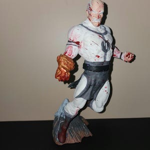 May include: A detailed figurine of a superhero in a dynamic pose. The figure is predominantly white with gray accents and red splatters, suggesting a battle scene. The right hand is a metallic gold color. The figure is mounted on a rocky base.