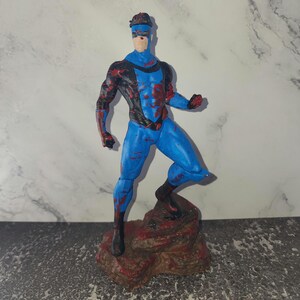 Può includere: Action figure di un supereroe blu e nero con dettagli rossi, in posa su una base rocciosa. La figura è dipinta con un body blu, finiture nere e dettagli rossi. La figura è alta circa 15 cm.