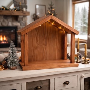 May include: A wooden creche structure with a gabled roof and an open front, featuring integrated warm LED string lights tucked under the roofline. The natural brown wood grain is visible throughout the frame, which stands on a rectangular base.