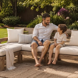 May include: An L-shaped outdoor sectional sofa made of weathered brown wood with thick off-white cushions. The set includes a chaise lounge section, a matching rectangular throw pillow, two square throw pillows, and a draped cream-colored throw blanket.