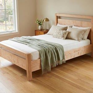 Op de afbeelding: Een lichtgekleurd houten bedframe met een hoofdbord en voetbord. Het bed is opgemaakt met een witte matras, kussens en een groene deken. De kamer heeft hardhouten vloeren en een raam.