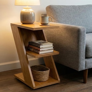 Puede incluir: Una mesa auxiliar de madera clara con un diseño único en forma de Z. Cuenta con dos estantes, uno con libros y otro con una cesta tejida. Una lámpara y una taza están encima. La mesa está junto a un sofá gris.