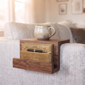 May include: A dark wood sofa arm table featuring a flat top surface and a vertical storage slot. The table holds a speckled ceramic mug on top, while two gold-coloured books and a black and gold pen are placed inside the integrated storage compartment.
