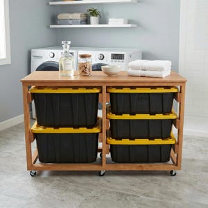 May include: A wooden storage cart with black and yellow storage bins. The cart has a wooden top with a glass bottle, a jar, and a bowl. White towels are stacked on the top. The cart is on wheels.